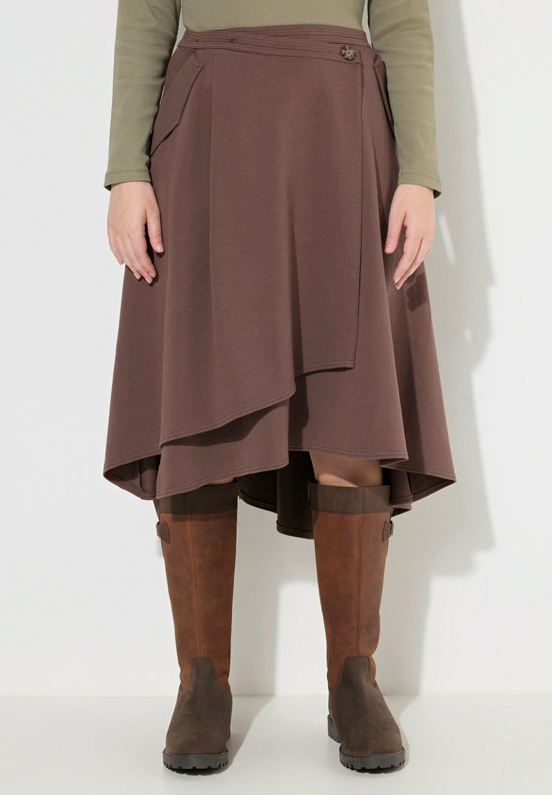 Bruine wikkelrok met een asymmetrische zoom, gemaakt van zacht materiaal; heeft zijzakken en een decoratieve knop. Gecombineerd met bruin-kneehoog laarzen.