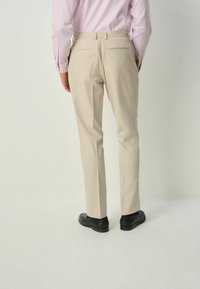 Man som bär beige skräddarsydda byxor, ljusrosa skjorta och svarta finskor, stående mot en ljus, enfärgad bakgrund.
