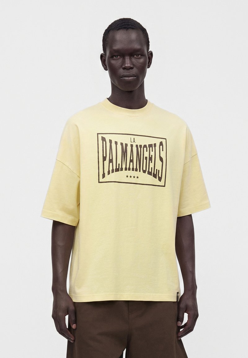 Man wearing a loose pale yellow T-shirt with "L.A. PALM ANGELS" text and four stars, paired with dark brown pants, standing against white background.