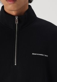 Sweatshirt zippé noir en tissu doux, avec un col haut et une fermeture éclair argentée. Logo blanc notable : "Abercrombie | NYC."