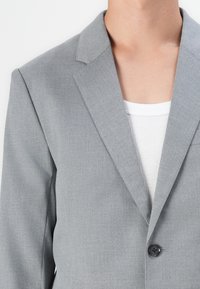 Light gray blazer featuring a notched lapel, single black button, and smooth fabric texture, worn over a white ribbed shirt.