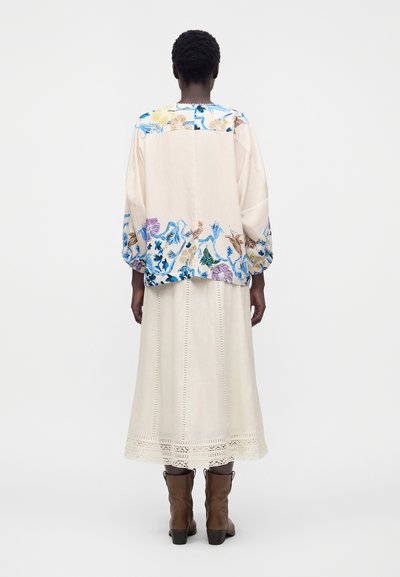 Personne portant un chemisier crème à fleurs avec des accents bleus, jaunes et violets, une jupe crème bordée de dentelle, et des bottes marron mi-mollet, de dos.