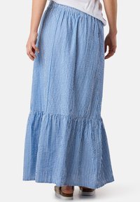 Jupe maxi rayée bleue et blanche avec un ourlet en couches, une taille élastique et un tissu texturé, portée avec des sandales brunes.