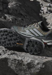 Utomhusvandrar skor med aggressiva svarta gummisulor, gröna och grå överdelar, vilande på klippig terräng under naturligt ljus.