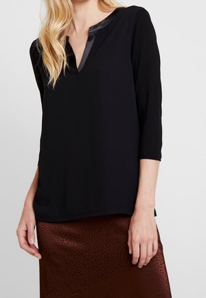 Woman with wavy blonde hair wears a black long-sleeve blouse with satin collar and brown patterned skirt, shown from shoulders to knees.