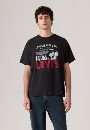 Ungdommelig mann ikledd en svart Levi's t-skjorte med hvit og rød tekst samt et grafisk motiv av en hest og rytter, kombinert med mørkeblå jeans.