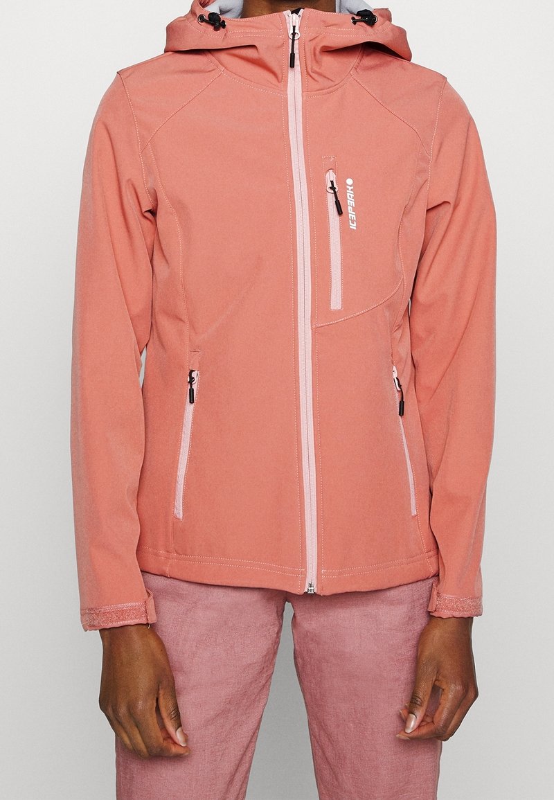 Personne portant une veste softshell rose avec fermeture éclair et capuche, et un pantalon rose assorti, debout devant un fond clair uni.