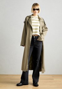 Lang oliven trench coat over en stripete crop top, kombinert med svarte slengjeans og svarte klumpete støvler. Tilbehør inkluderer mørke solbriller.