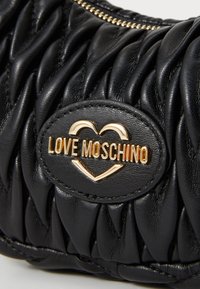 Mustmust nahk käekott, millel on kuldne logo patch, millel on kiri "LOVE MOSCHINO" ja eriline südame disain. Lukuga sulgemine.