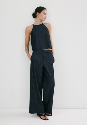 Femeie stând lângă un perete alb, purtând un top fără mâneci de culoare albastru închis, pantaloni largi albastru închis și pantofi negri cu vârf deschis.