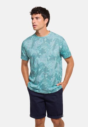 Hombre con una camisa de manga corta color verde azulado con estampados de hojas y pantalones cortos azul marino remangados, de pie con las manos en los bolsillos mirando hacia la izquierda.