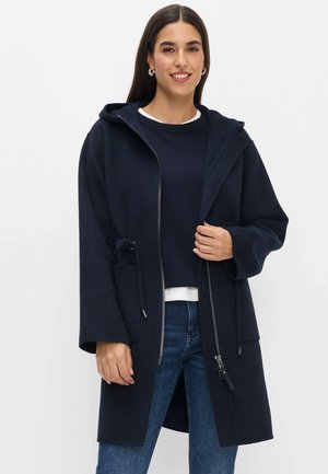 Kvinde iført en mørkeblå hættejakke over en mørkeblå sweater og blå jeans, stående foran en ensfarvet hvid baggrund.