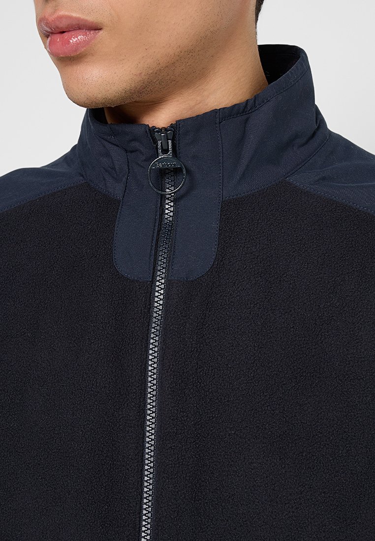 Veste en polaire noire avec col montant et fermeture éclair. Caractérisée par un corps texturé et des sections supérieures lisses. Tirette de fermeture éclair avec un accent en anneau.