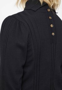 Vêtement noir en tricot avec un col haut, texture côtelée verticale et cinq boutons marron le long du dos. Manches bouffantes visibles.