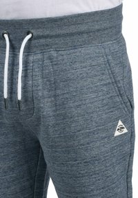 Blaugraue Sweatpants mit geripptem Bund und weißen Kordeln, ausgestattet mit Seitentaschen und einem dreieckigen Logo-Patch auf dem rechten Oberschenkel.