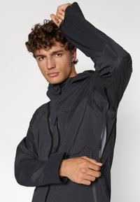 Veste noire imperméable avec capuche, dotée de poches zippées, de poignets ajustables et d'une texture mate lisse. Logo affiché sur la poitrine.