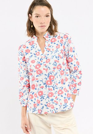 Femme portant un chemisier blanc à imprimé floral rose et bleu, manches trois-quarts, et pantalon beige, debout devant un fond uni.