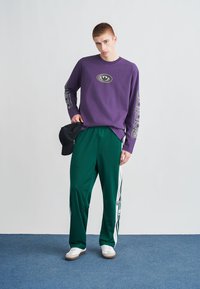 Violeta garām piedurknēm krekls ar centrālo logo, kopā ar zaļām sporta biksēm ar baltām sānu svītrām. Modelis valkā baltas sporta kurpes un cepuri.