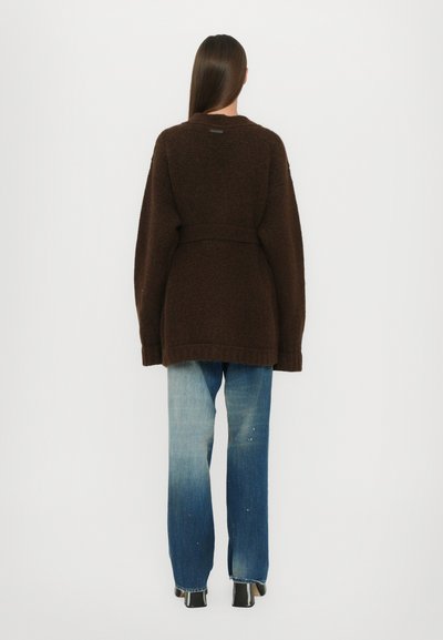 Brown knitted cardigan with a belted waist. Features a relaxed fit, long sleeves, and a textured finish. Worn with light-wash jeans.