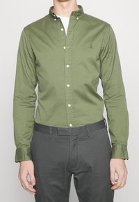 Grön bomullsskjorta med knappar och krage, långa ärmar och vita knappar. Har en liten logotyp på bröstet. Kombinerad med grå byxor.