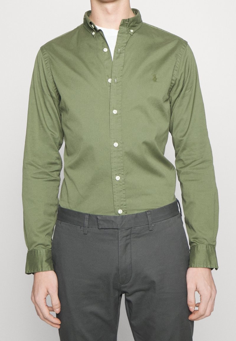 Grön bomullsskjorta med knappar och krage, långa ärmar och vita knappar. Har en liten logotyp på bröstet. Kombinerad med grå byxor.