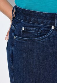 Tmavě modré džíny s vysokým pasem, stříbrnými kovovými detaily a malou přední kapsou. Materiál má hladkou strukturu.