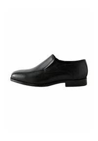 Zapato negro de cuero sin cordones con textura suave, puntera redondeada y detalles de costura mínimos. La suela baja y plana realza un diseño elegante.