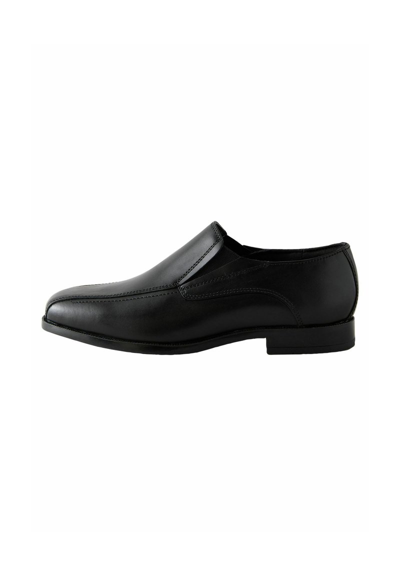 Zapato negro de cuero sin cordones con textura suave, puntera redondeada y detalles de costura mínimos. La suela baja y plana realza un diseño elegante.