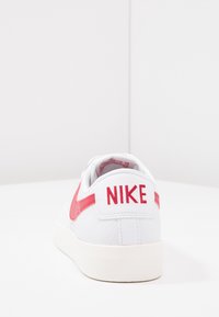 Vita sneakers med en texturerad läderöverdel, som har en röd Nike swoosh och logotyp på hälkappan, samt en vit gummisula.