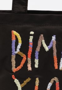 Czarna torba na ramię z tkaniny, ozdobiona kolorowym haftowanym napisem z różnorodnymi fakturami nici w odcieniach pomarańczowym, fioletowym, żółtym i brązowym.