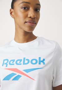 Balta medvilninė marškinėliai su dideliu mėlynu "Reebok" logotipu ir raudonu akcentų raštu, trumpomis rankovėmis, apvaliu kaklu ir glotnia tekstūra.