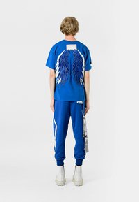 Camiseta azul con detalle gráfico de alas, combinada con joggers azules que presentan acentos en blanco y una etiqueta lateral. El modelo lleva botas blancas.