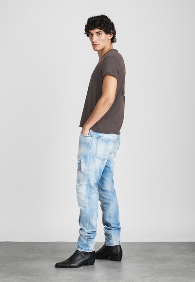 T-shirt castanho de manga curta, jeans azuis claros desgastados e botas pretas até ao tornozelo. Modelo está de lado contra um fundo cinza.