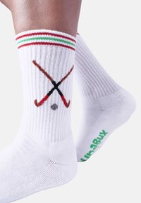 Calzini bianchi di cotone con trama a coste, con bordi a righe rosse, verdi e rosse, e grafica di una stecca da hockey con disco. Logo verde sulla suola.