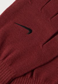 Červené pletené rukavice s textúrovaným vzorom a čiernym logom Nike na zápästí. Mäkký materiál a priliehavý strih.