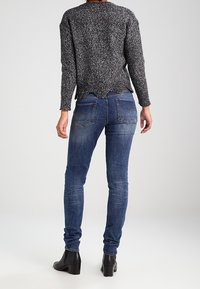 Suéter de punto gris con textura y dobladillo en forma de ondas, combinado con jeans ajustados azul oscuro y botas de tobillo negras. Vista trasera que muestra los detalles de la ropa.