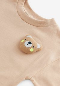 Helles beiges Sweatshirt mit einer bärenförmigen Stoffapplikation, gelben Wangenakzenten und einer weißen Gesichtsillustration. Sanfte Textur.