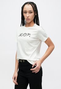 Λευκό κοντομάνικο T-shirt με μαύρο γραφικό "JOOP!", συνδυασμένο με μαύρο παντελόνι. Απαλό βαμβακερό ύφασμα, άνετη εφαρμογή, χωρίς σχέδια.