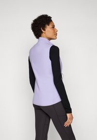 Vest lila cu guler înalt, închidere cu fermoar și design ajustat, asortat cu un top negru cu mânecă lungă și pantaloni maro închis. Textură netedă.