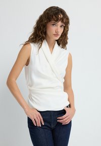 Hvid ærmeløs top med draperet halsudskæring og indpakningsdesign, lavet af et glat stof; parret med mørke denimjeans.