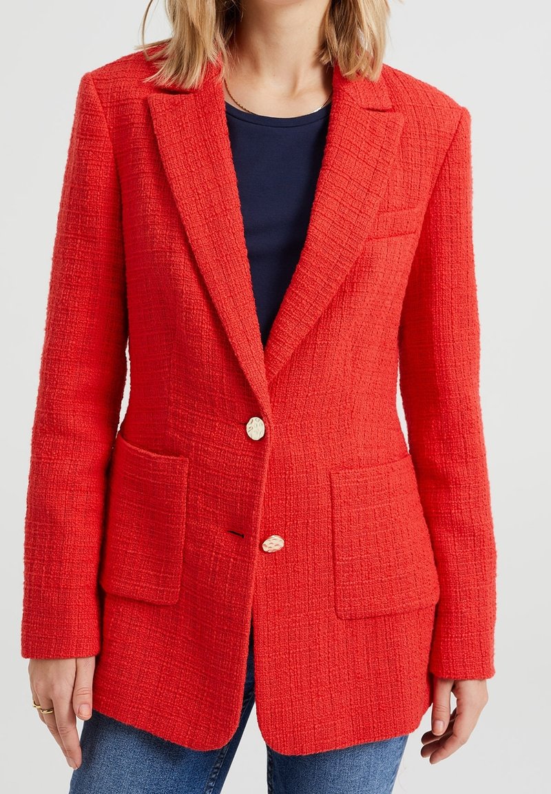 Femme portant un blazer rouge texturé avec deux poches avant et des boutons dorés sur un top bleu marine et un jean bleu.