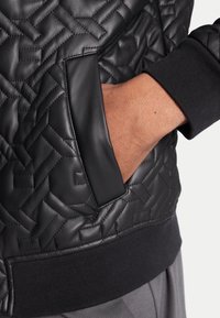 Chaqueta de cuero negra acolchada con un bolsillo lateral con un acento de cuero suave y puños acanalados, que exhibe un patrón texturizado.
