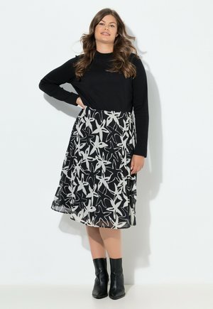 Woman with wavy hair wearing a black long-sleeve top, a black and white floral patterned skirt, and black ankle boots standing against a wall.