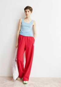 Blaues ärmelloses Tanktop kombiniert mit lockeren roten Jogginghosen mit weißem Logo, dazu eine hellviolette Tragetasche und weiße Sneaker.