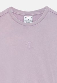 T-shirt violet clair avec un col rond côtelé et un petit logo "C" brodé sur la poitrine. Texture de tissu douce et design décontracté.
