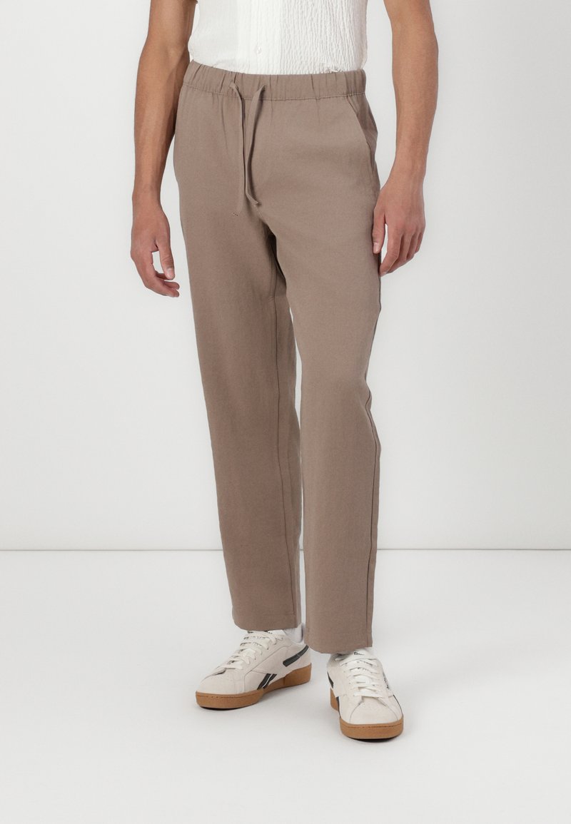 Beige, leichte, lässige Hose mit elastischem Bund und Zugband, kombiniert mit weißen Sneakers vor weißem Hintergrund.