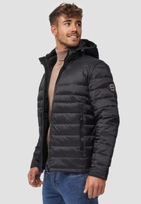 Veste noire matelassée avec une capuche, présentant une texture en quilt, des zips sur les côtés et un patch logo sur la manche gauche. Portée par-dessus un léger col roulé.
