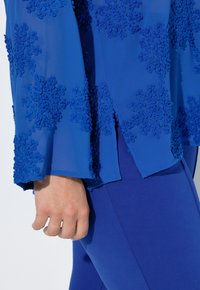 Blå gennemsigtig top med tekstureret, blomsterbroderi. Har vide ærmer og lagdelt kant. Kombineret med ensfarvede, blå tætsiddende bukser.