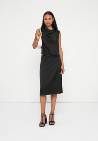 Schwarzes, ärmelloses Oberteil mit drapiertem Ausschnitt, kombiniert mit einem taillierten schwarzen Bleistiftrock. Das Modell hält eine hellrosa Handtasche. Strukturierter Stoff, unaufdringliches Design.