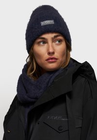 Marine gebreide beanie met een textuurafwerking en een logopatch, gecombineerd met een bijpassende sjaal en een zwarte waterdichte jas met opvallende hardware.
